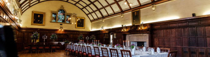 Large dining hall with oak panelled walls, stained glass windows, historic paintings. A long table sits down the centre dressed for dining. Piano is just under the camera at the front.